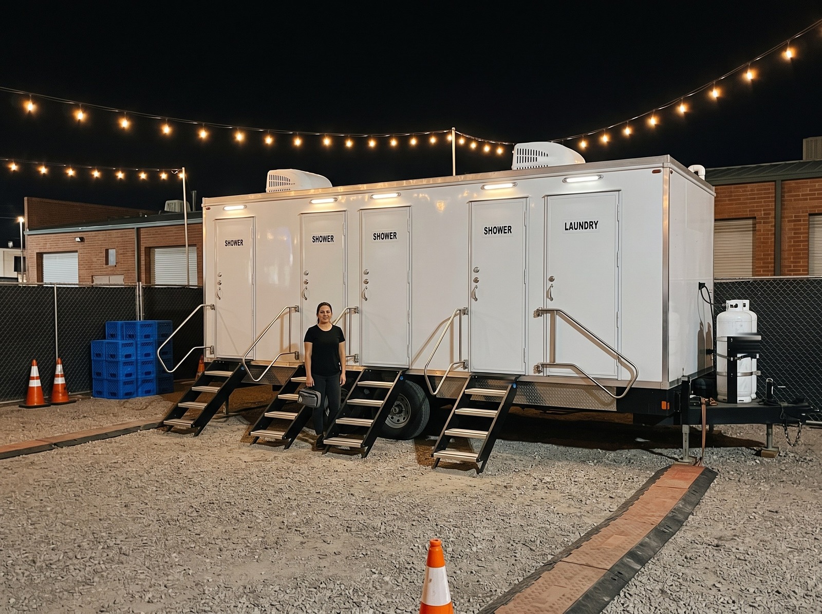 Four station shower and laundry trailer lit up at night in a fenced gravel event lot under strings of bulbs